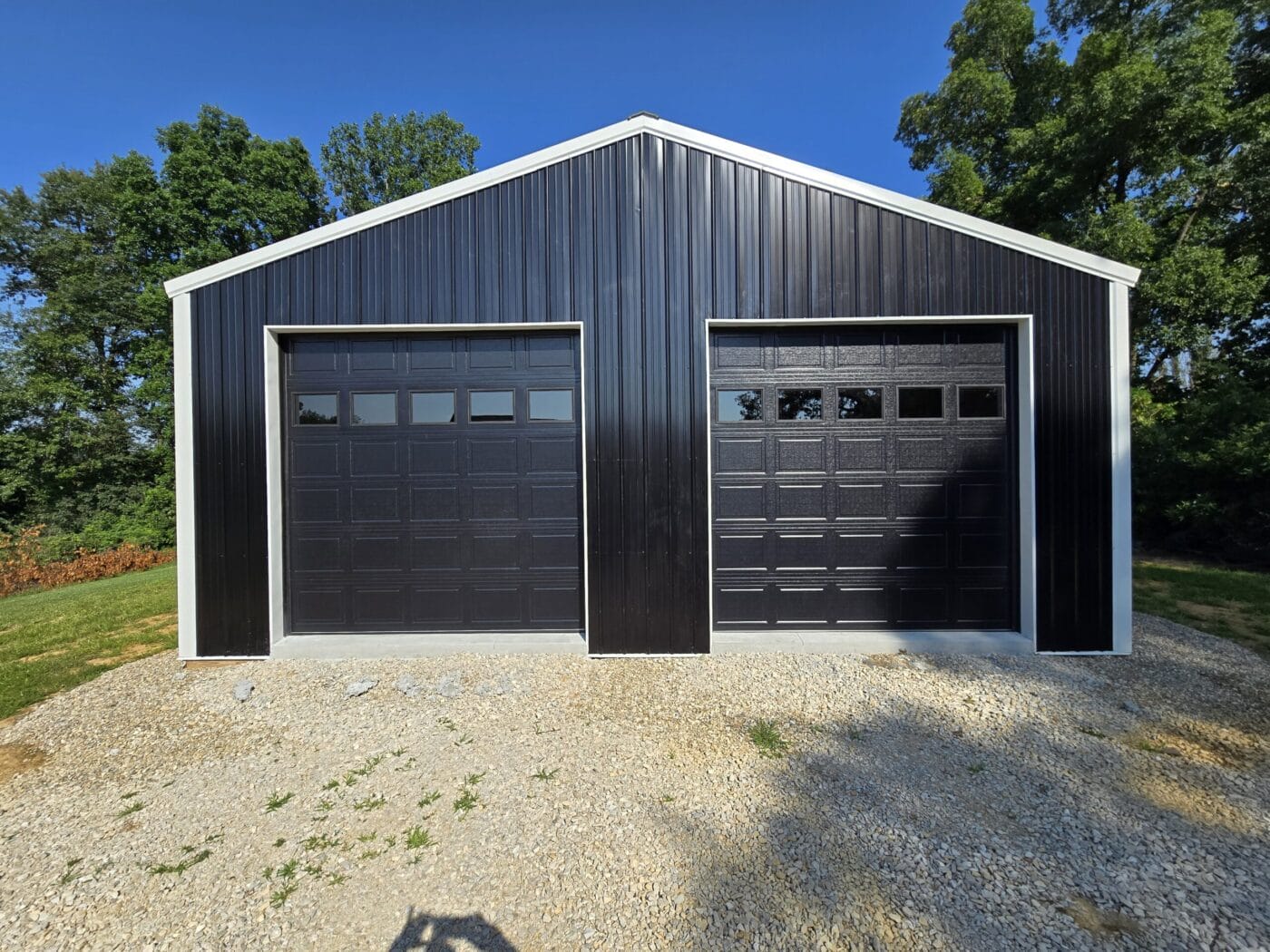 A black metal building with two large garage doors, surrounded by gravel and grass, set against a backdrop of trees under a clear blue sky.