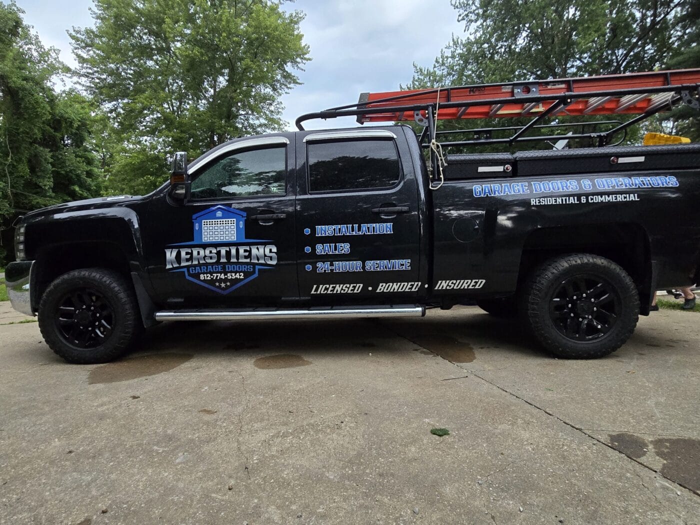 A black truck with Kerstiens Garage Doors branding on the side, parked on a driveway. A red ladder is secured on top.