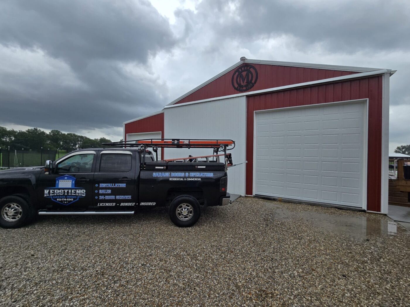 A truck with ladder racks is parked outside a large red and white warehouse-style building on a gravel driveway under a cloudy sky.