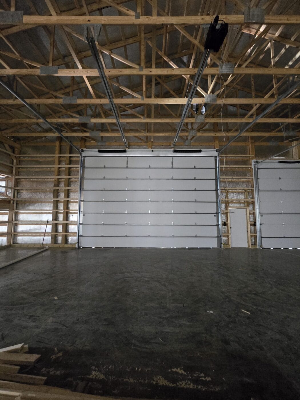 Interior of a spacious, unfinished garage with a high ceiling, exposed wooden beams, and large white garage doors.