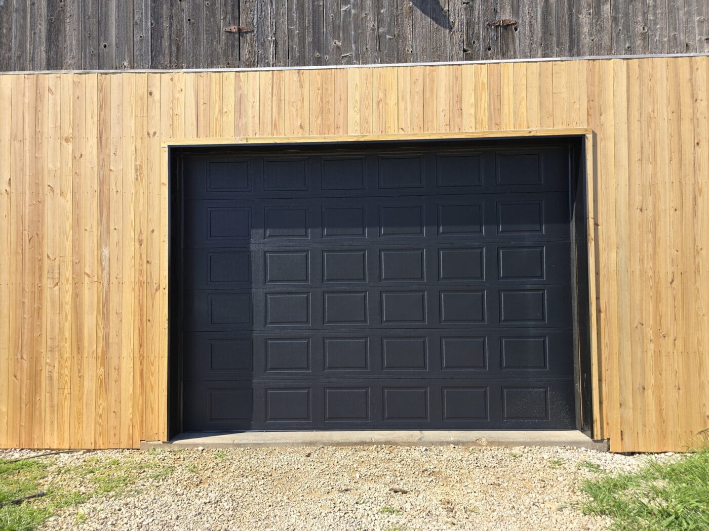 A black garage door encased in light wood paneling, with gravel on the ground.