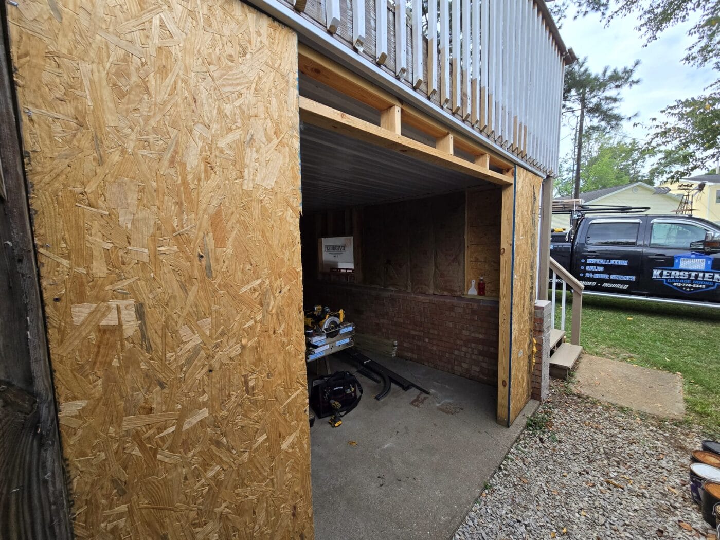 A partially constructed or renovated garage with exposed wooden panels and construction tools inside. A branded vehicle is parked outside on a gravel driveway.