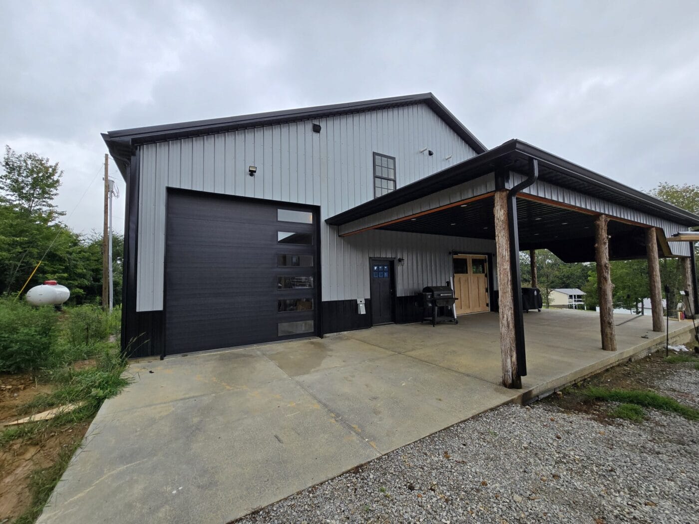 A large gray metal building with a black garage door and a covered porch supported by wooden beams. Overcast sky in the background.