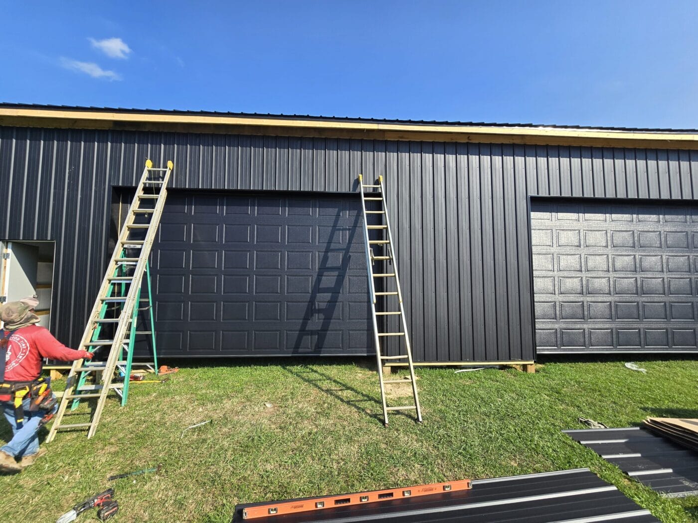 Construction workers position ladders against a black metal building with garage doors on a sunny day.