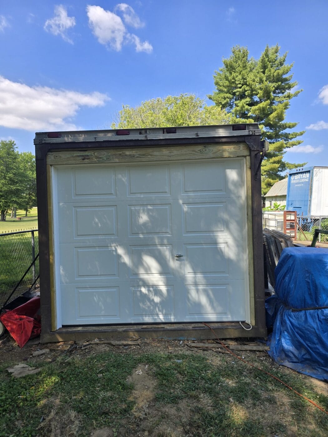 A white garage door is installed within a wooden frame outside, surrounded by grass and various objects, under a blue sky with scattered clouds.