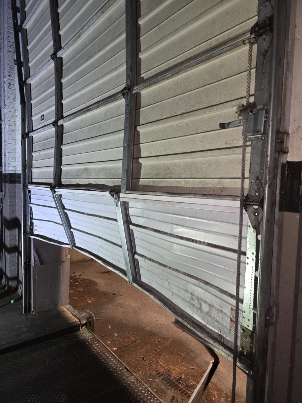 A damaged industrial garage door with a bent section is partially open, exposing a dimly lit area outside.