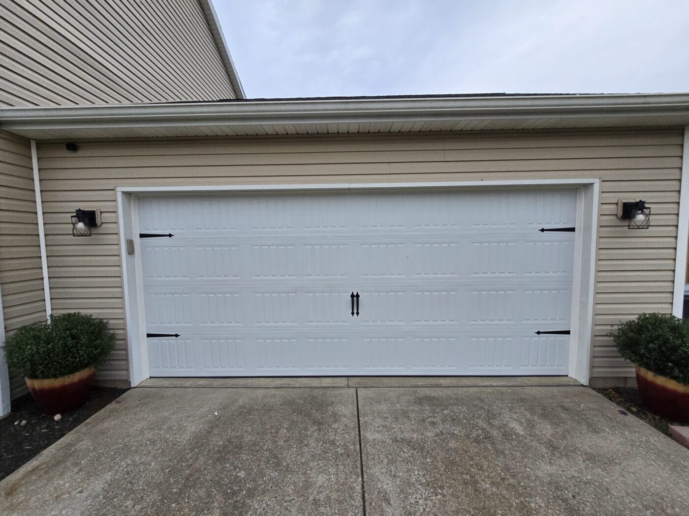 A white garage door with black handles is flanked by two potted plants and lantern-style lights on either side. It is set in a beige siding exterior.