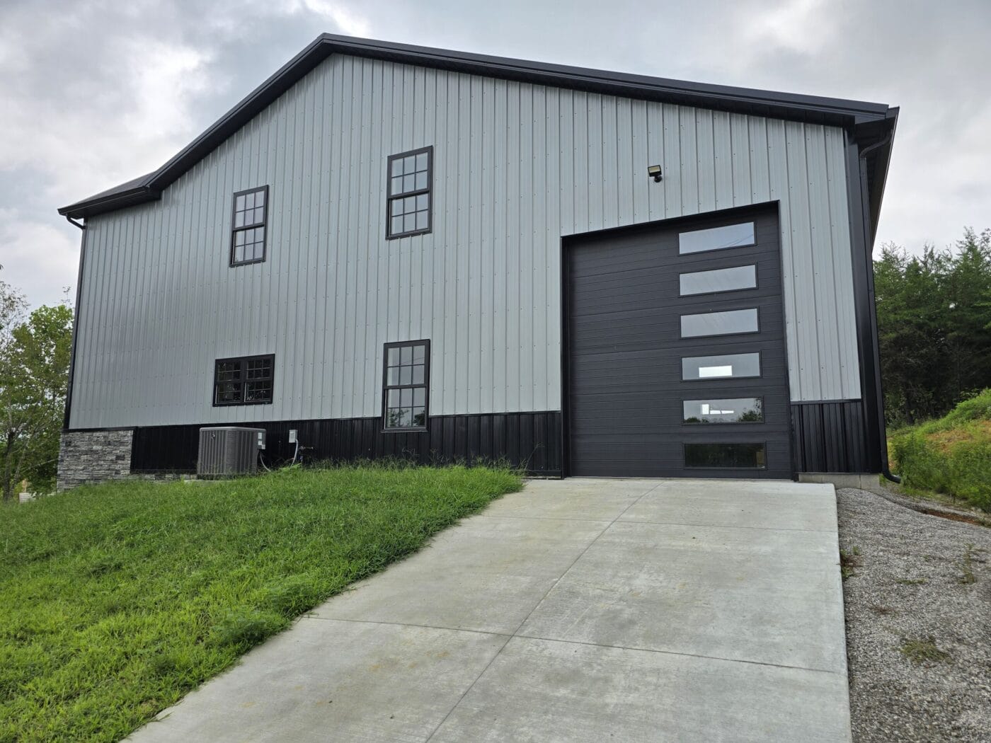 A gray metal building with a large black garage door and several windows, set on a grassy slope with a concrete driveway.